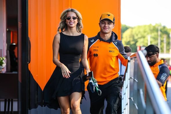 Margarida Corceiro dalam gaun hitam dan Lando Norris dengan hoodie oranye dan celana gelap berjalan ke paddock saat balapan